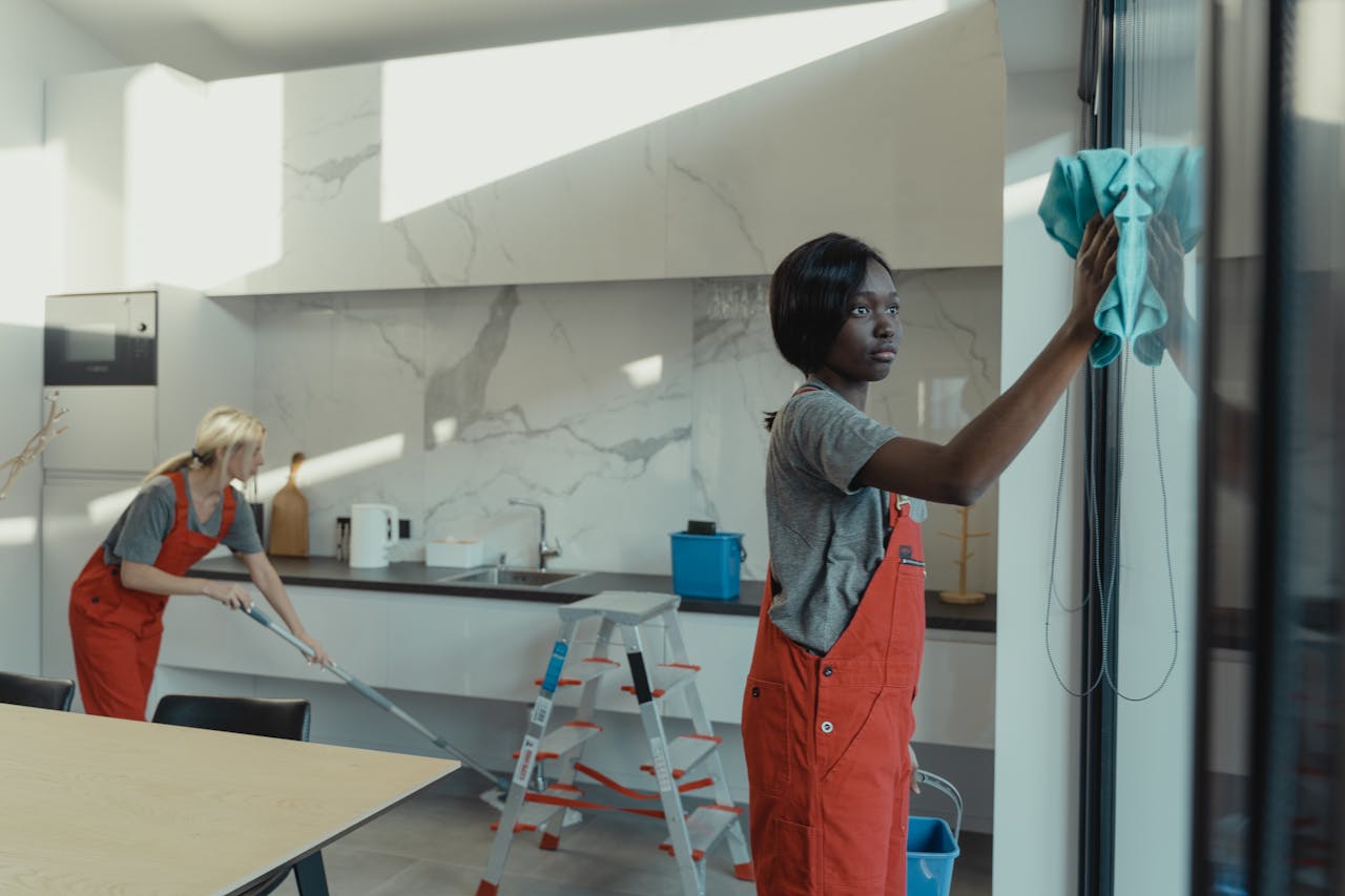 hero-img Two women cleaning a modern kitchen, wiping glass surfaces and mopping the floor.